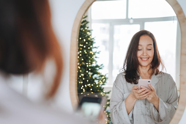 Junge Erwachsene, vierzig Jahre, schöne Frau mittleren Alters mit dunklen Haaren in gemütlichen Pyjamas, die in den Spiegel schauen und am Weihnachtsmorgen im Zimmer des Hauses ein Selfie machen