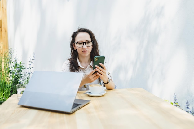 Junge Europäerin sitzt mit Laptop und einer Tasse Kaffee am Tisch im Freien, lächelt Frau mit Brille und genießt die Telearbeit im Café oder das Online-Lernen