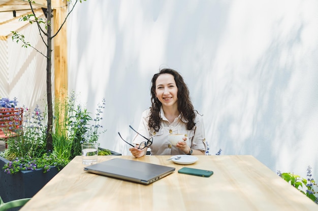 Junge Europäerin sitzt mit Laptop und einer Tasse Kaffee am Tisch im Freien, lächelt Frau mit Brille und genießt die Telearbeit im Café oder das Online-Lernen