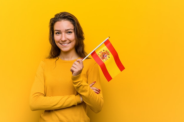 Junge europäische Frau, die ein spanisches Flaggenlächeln überzeugt mit den gekreuzten Armen hält.