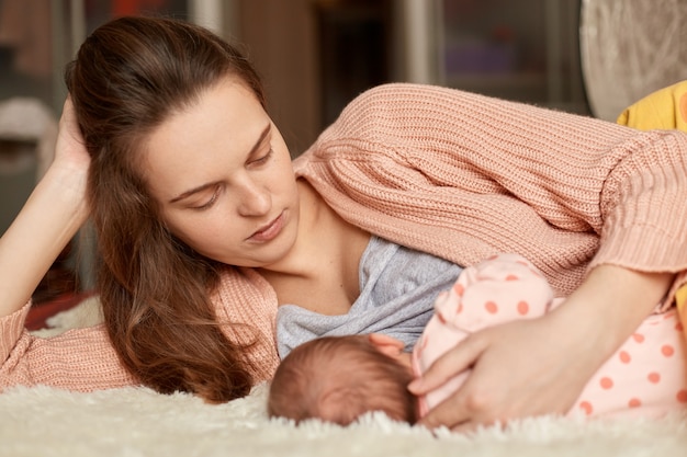 Junge europäische Frau, die Hauskleidung trägt, die im Bett mit ihrem neugeborenen Baby liegt, Säugling betrachtet und füttert, kleines Kind isst, stillt, Mama mit Kind zu Hause