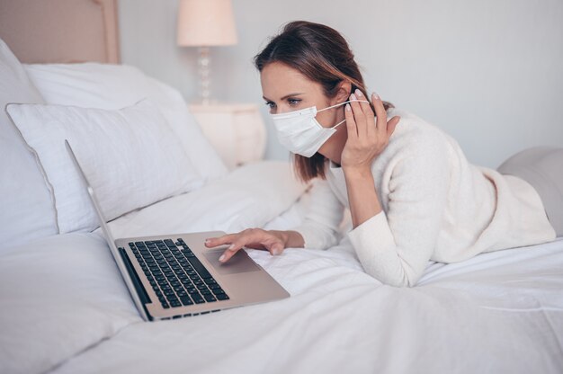 Junge europäische Frau in Gesichtsmedizinmaske, die an einem Laptop im Schlafzimmer während der Coronavirus-Isolierung arbeitet