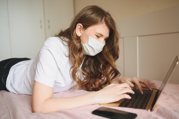 Junge europäische Frau in Gesichtsmedizinmaske, die an einem Laptop und Telefon im Bett arbeitet