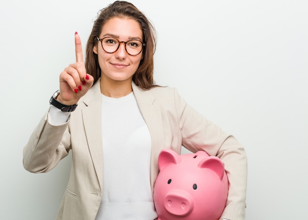 Junge europäische Geschäftsfrau, die ein Sparschwein zeigt Nummer Eins mit dem Finger hält.