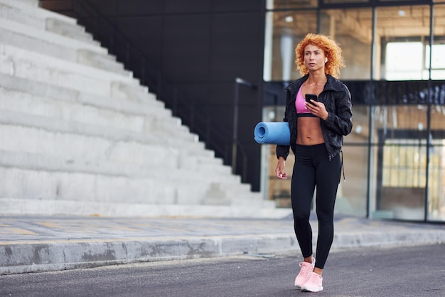 Foto junge europäische rothaarige frau in sportlicher kleidung mit telefon und matte in den händen im freien