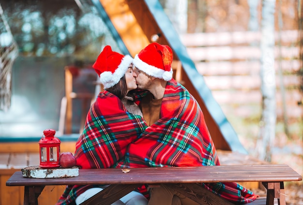 Junge Familie in der Weihnachtsmütze, die auf dem hölzernen alten Tischhintergrund ihres Hauses sitzt