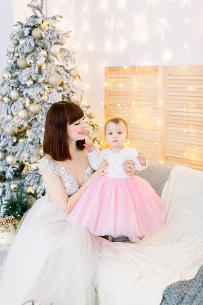 Junge Familie, Mutter mit Baby, sitzend auf dem Sofa, auf dem Hintergrund des Weihnachtsbaumes