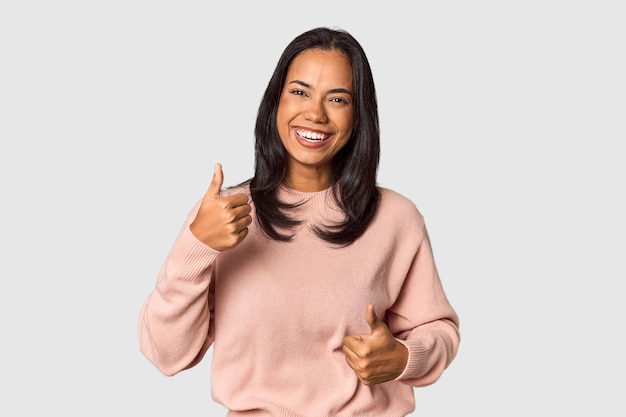 Junge Filipina mit langen schwarzen Haaren im Studio, die beide Daumen hochhebt, lächelnd und zuversichtlich