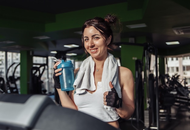 Junge fit Frau mit einem Handtuch auf ihren Schultern, das Wasserflasche auf einem Laufband hält. Gewichtsverlust Konzept, Aerobic Workout