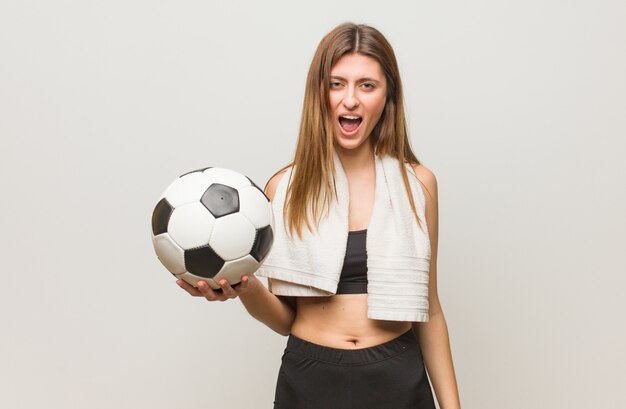 Junge Fitness russische Frau, die sehr wütend und aggressiv schreit. Einen Fußball halten.