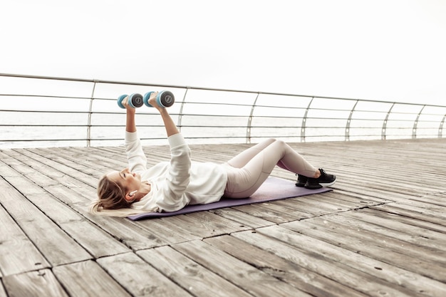 Junge Fitnessfrau, die im Freien auf der Matte liegend Hantelpresse übt. Gesunder Lebensstil