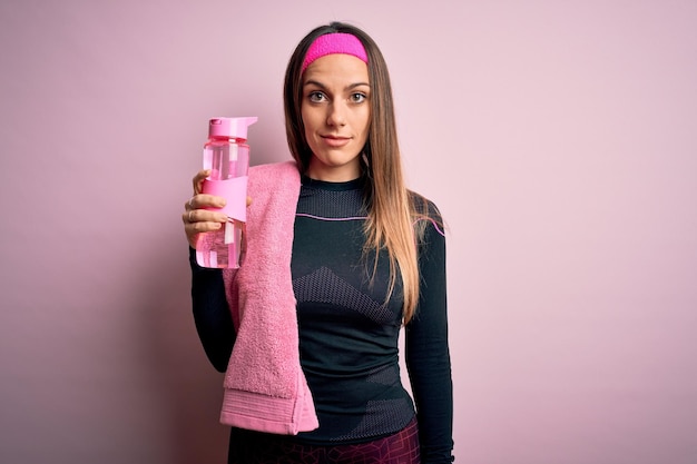 Junge Fitnessfrau, die Sportkleidung und Handtuchtrinkwasser aus der Flasche zum Mitnehmen trägt, mit einem selbstbewussten Ausdruck auf einem intelligenten Gesicht, das ernsthaft denkt