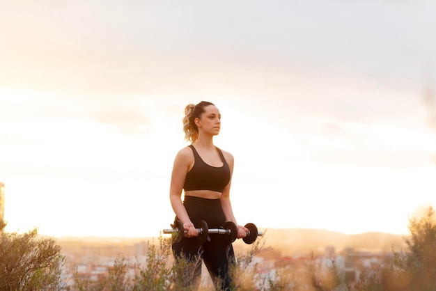 Junge Fitnessfrau hebt Gewichte im Freien bei Sonnenuntergang