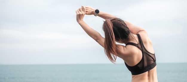 Junge Fitnessfrau in Sportkleidung, die Körper gegen Meerblick dehnt, gesunde Frauenübung am Morgen. Trainings-, Wellness- und Work-Life-Balance-Konzepte