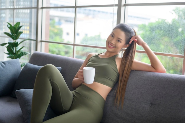 Junge Fitnessfrau in Sportkleidung, die nach dem Training eine Tasse Kaffee trinkt