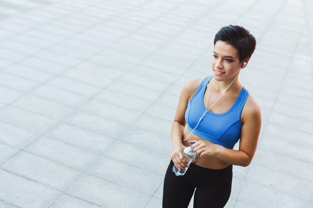 Junge Fitnessfrau mit Kopfhörern macht Pause, trinkt Wasser nach dem Training in der Stadt, schaut weg und lächelt, kopiert Platz