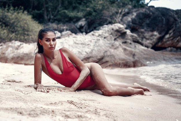 Junge, fitte Frau im roten Badeanzug, die am nassen Sandstrand liegt, mit unscharfen Felsen im Hintergrund. Freiheitsstrand, Phuket