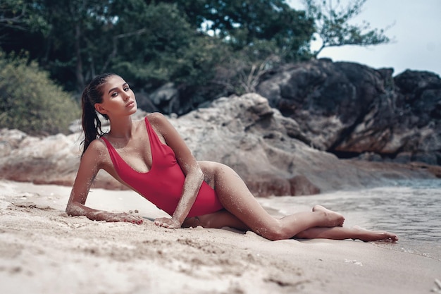 Junge, fitte Frau im roten Badeanzug, die am nassen Sandstrand liegt, mit unscharfen Felsen im Hintergrund. Freiheitsstrand, Phuket