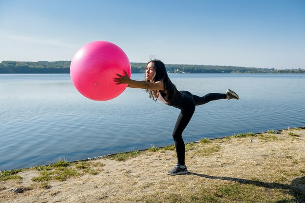 Junge flexible Frau, die Morgenfitnessübungen auf rosa Ball nahe See macht