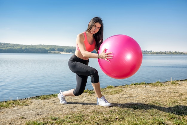 Junge flexible Frau macht morgendliche Fitnessübungen auf rosa Ball in der Nähe des Sees