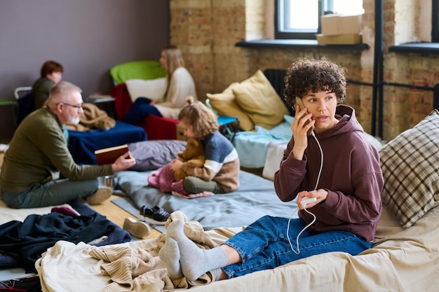 Junge Flüchtlingsfrau, die per Smartphone anruft, während sie auf einem Schlafplatz sitzt