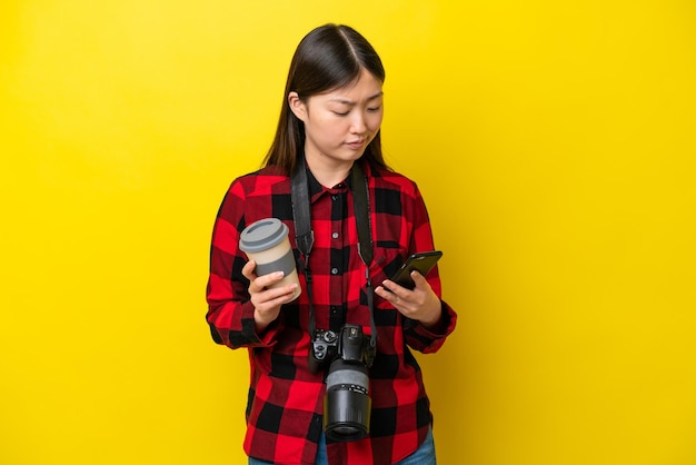 Junge Fotografin Chinesin isoliert auf gelbem Hintergrund mit Kaffee zum Mitnehmen und einem Handy