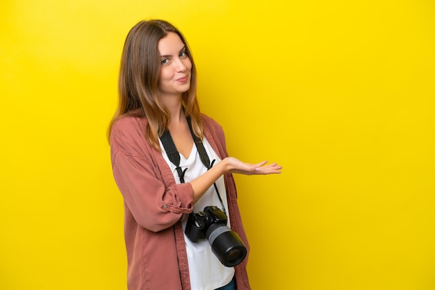 Junge Fotografin, kaukasische Frau isoliert auf gelbem Hintergrund, die eine Idee präsentiert, während sie lächelnd in Richtung schaut