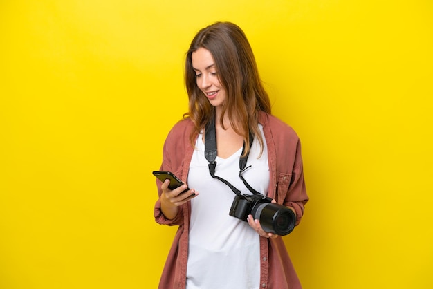 Junge Fotografin, kaukasische Frau isoliert auf gelbem Hintergrund, die eine Nachricht mit dem Handy sendet