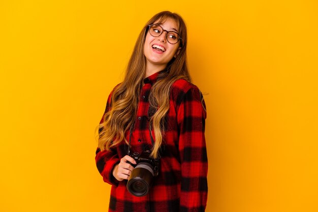 Junge Fotografin lokalisiert auf gelber Wand schaut lächelnd, fröhlich und angenehm beiseite