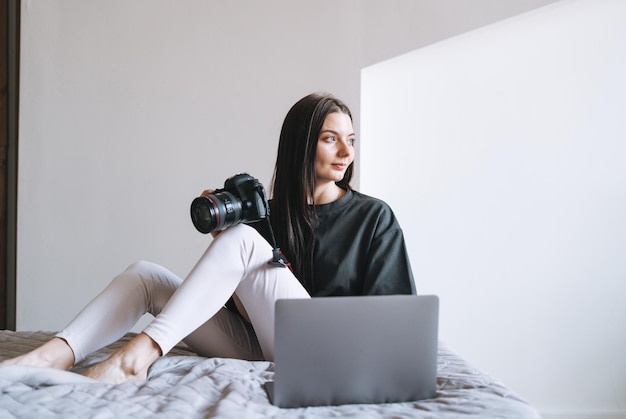 Junge Fotografin mit Digitalkamera-Freiberuflerin in Freizeitkleidung mit Laptop, der zu Hause auf dem Bett sitzt