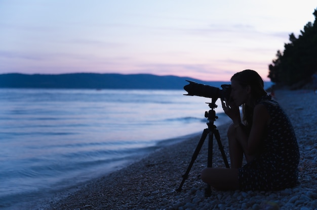 Junge Fotografin sitzt am Kiesstrand am Abend mit ihrer Kamera auf einem Stativ, bereit, das abendliche Meer und die Landschaft zu fotografieren.