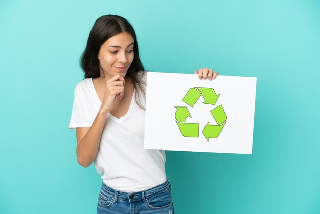 Junge französische Frau lokalisiert auf blauem Hintergrund, der ein Plakat mit Recycling-Symbol hält