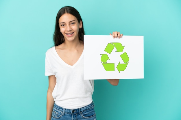 Junge französische Frau lokalisiert auf blauem Hintergrund, der ein Plakat mit Recycling-Symbol mit glücklichem Ausdruck hält