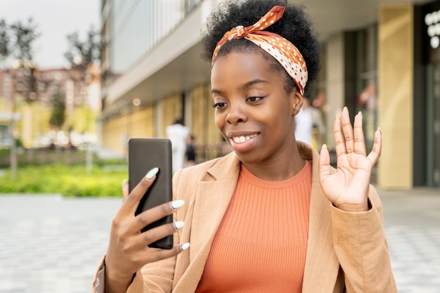 Junge Frau afrikanischer Abstammung, die ihren Freund auf dem Smartphone-Display mit einem zahnigen Lächeln und einer winkenden Hand während des Gesprächs ansieht