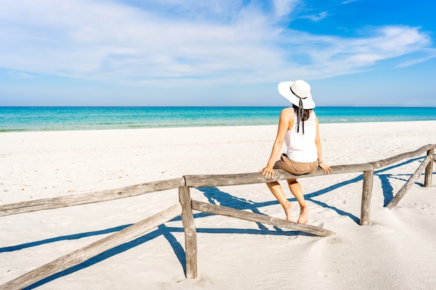 Junge Frau allein im Urlaub bewundert das kristallklare tropische Meer, das auf einem Holzzaun an einem weißen Sandstrand unter blauem Himmel sitzt. Nachdenkliches Mädchen mit großem weißen Hut genießt Sommerreisen