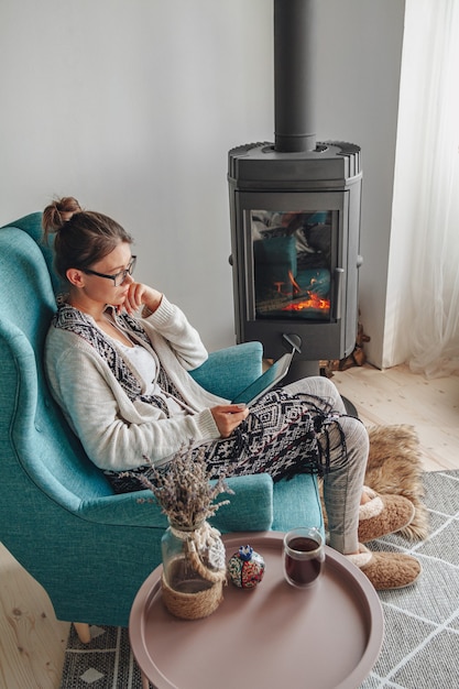 Junge Frau am Kamin, sitzend in einem gemütlichen Sessel, mit einer warmen Decke, mit einer Tablette