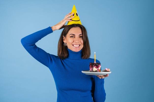 Junge Frau an blauer Wand feiert Geburtstag, hält ein Stück Kuchen, gut gelaunt, glücklich, aufgeregt
