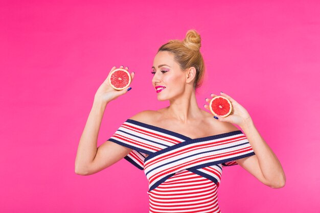 Junge frau an einer rosa wand hält eine geschnittene orange in ihren händen und lacht.