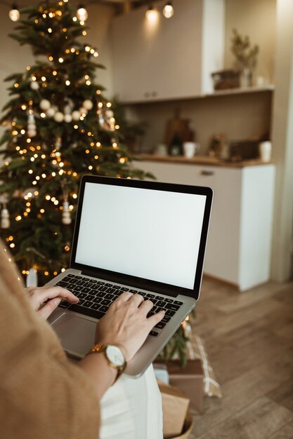 Junge Frau arbeiten am Laptop-Computer mit leerem Bildschirm mit Kopierraum. Modernes Hauptwohnzimmerinterieur, das für Weihnachtsfeier mit Weihnachtsbaum- und Girlandenlichtern verziert wird