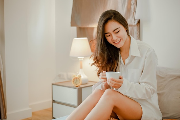 Junge Frau Asien, die zu Hause lebt, sich entspannt und im Urlaub eine Tasse heißen Kaffee im Schlafzimmer trinkt. Asiatisch, Asien, Entspannung, allein, Technologie, Lifestyle-Konzept.