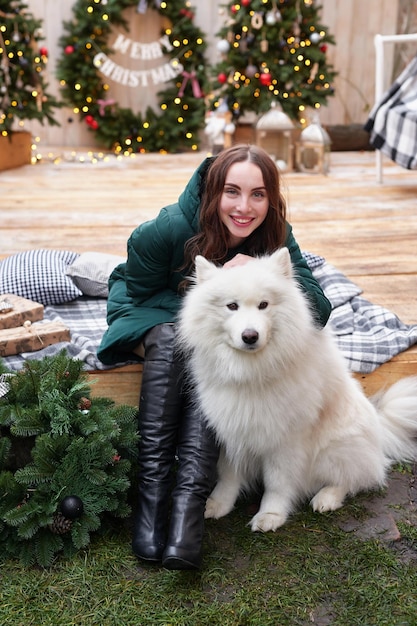 Junge Frau auf dem Hintergrund des Weihnachtsbaums mit weißem Samoyed-Hund im Freien Hofdekoration für das neue Jahr