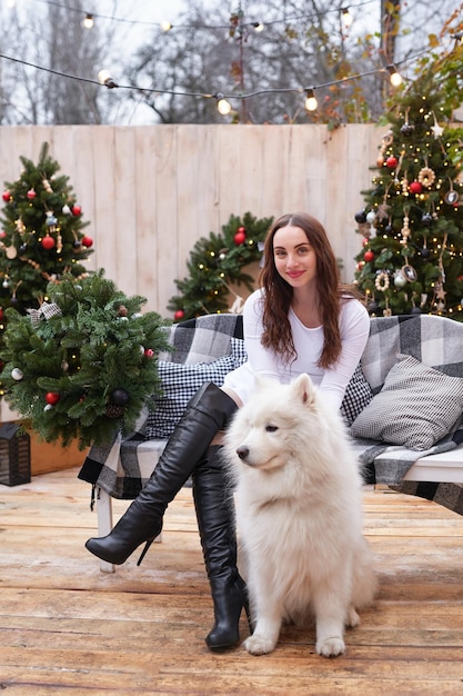 Junge Frau auf dem Hintergrund des Weihnachtsbaums mit weißem Samoyed-Hund im Freien Hofdekoration für das neue Jahr