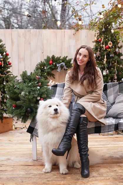 Junge Frau auf dem Hintergrund des Weihnachtsbaums mit weißem Samoyed-Hund im Freien Hofdekoration für das neue Jahr