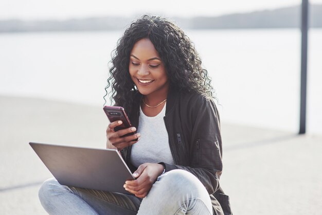 Junge Frau auf der Straße, die am Laptop arbeitet und auf Handy spricht