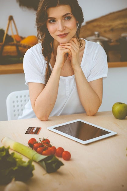 Foto junge frau auf der suche nach einem neuen rezept zum kochen in einer küche. hausfrau macht online-shopping per tablet-computer und kreditkarte.