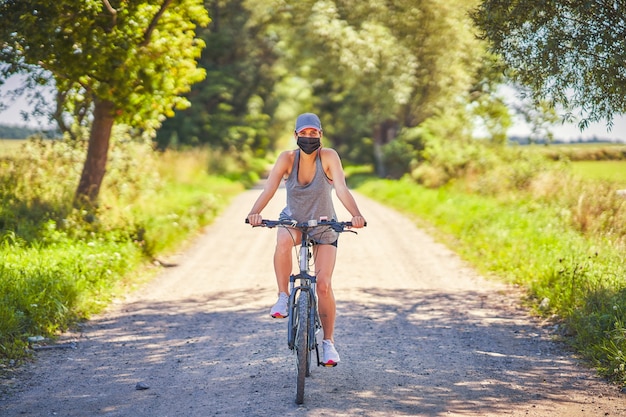 Junge Frau auf einem Fahrrad auf dem Land, die aufgrund einer Coronavirus-Pandemie eine Maske trägt