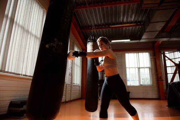 Junge Frau beim Boxtraining im Fitnessstudio, sie trägt Boxhandschuhe und schlägt einen Boxsack.