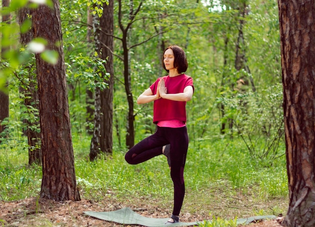 Junge Frau beim Yoga im grünen Sommerwald Frau, die in Vrikshasana zwischen den Bäumen steht