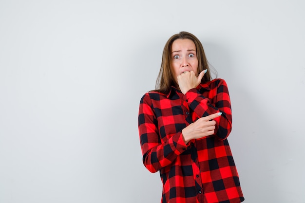 Junge Frau beißt Faust, mit der Hand auf dem Bauch, zeigt mit dem Zeigefinger im karierten Hemd nach rechts und sieht besorgt aus, Vorderansicht.