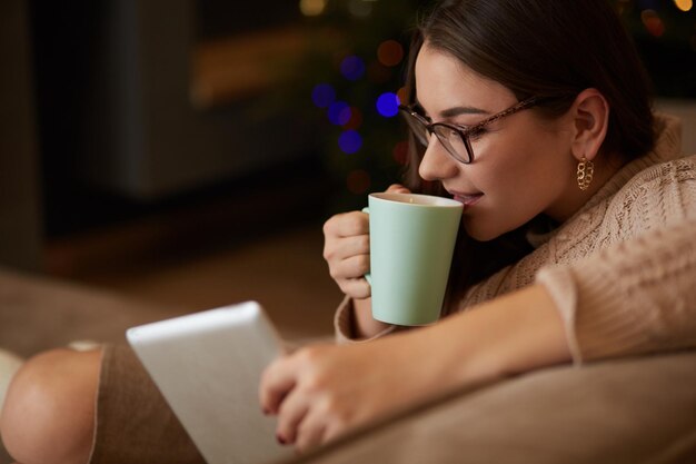 Junge Frau benutzt Tablet, während sie sich zu Hause mit Kaffee auf der Sofacouch entspannt